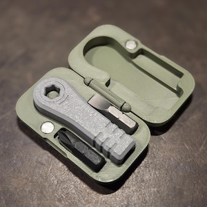 May include: An open, olive-green, rectangular case with a gray multi-tool set. The set includes a wrench, a flathead screwdriver, and a Phillips head screwdriver. The case has a carabiner-style clip.