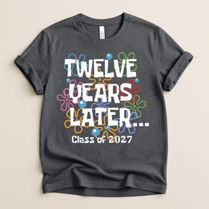 May include: A dark gray short-sleeve t-shirt featuring the white text TWELVE YEARS LATER... and Class of 2027. The design includes colorful, outlined flower shapes in blue, orange, pink, and green, along with small blue circles scattered throughout.