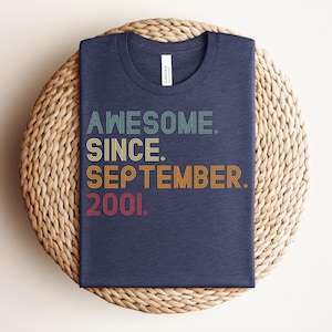 May include: A dark blue crew neck t-shirt featuring the text AWESOME. SINCE. SEPTEMBER. 2001. printed in a multi-line, retro-inspired font with horizontal line details in shades of teal, yellow, orange, and red against the fabric.