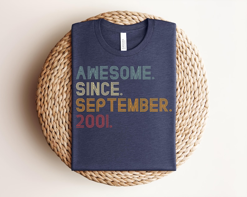May include: A dark blue crew neck t-shirt featuring the text AWESOME. SINCE. SEPTEMBER. 2001. printed in a multi-line, retro-inspired font with horizontal line details in shades of teal, yellow, orange, and red against the fabric.