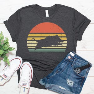 May include: A dark charcoal gray t-shirt featuring a distressed graphic of a black wild boar silhouette centered over a retro-style sunset design with horizontal stripes in shades of orange, yellow, and muted green.