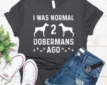 May include: A dark charcoal gray short-sleeve t-shirt featuring the white text I WAS NORMAL 2 DOBERMANS AGO with silhouettes of two Doberman dogs and small paw print graphics. The shirt is displayed alongside white canvas sneakers and blue denim shorts.