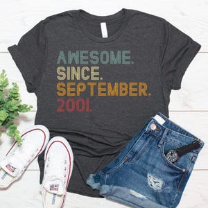 May include: A dark charcoal gray short-sleeve t-shirt featuring the text "AWESOME. SINCE. SEPTEMBER. 2001." printed in a retro-style font with stacked lines in shades of teal, yellow, orange, and red.