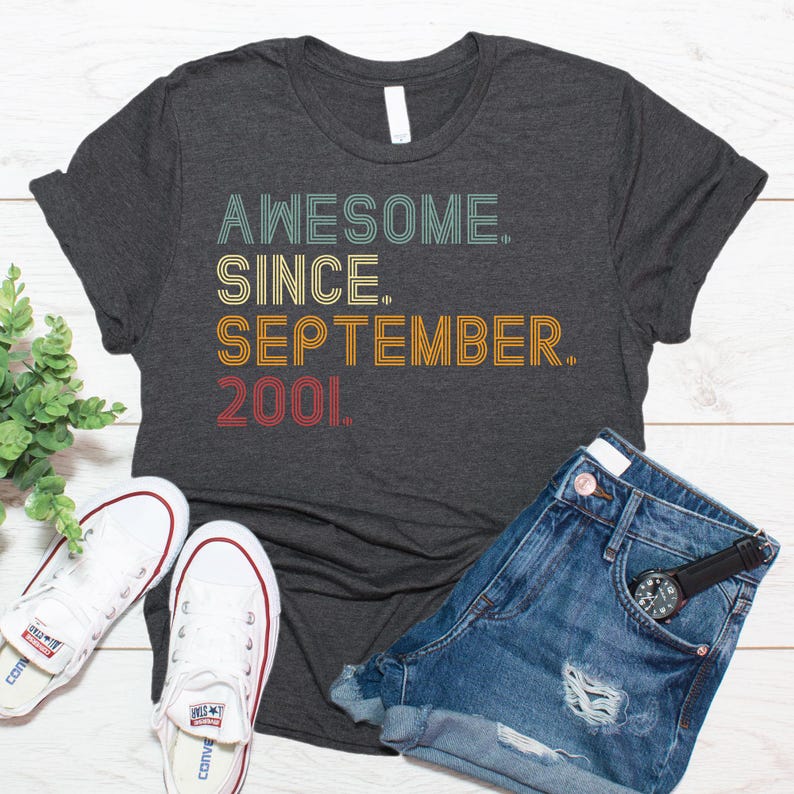 May include: A dark charcoal gray short-sleeve t-shirt featuring the text "AWESOME. SINCE. SEPTEMBER. 2001." printed in a retro-style font with stacked lines in shades of teal, yellow, orange, and red.
