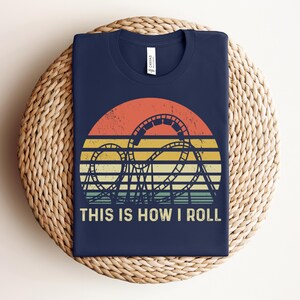 Może przedstawiać: Granatowa koszulka z nadrukiem w stylu retro z zachodem słońca i sylwetką kolejki g&oacute;rskiej. Pod grafiką znajduje się napis "THIS IS HOW I ROLL". Koszulka wykonana jest z miękkiego materiału.