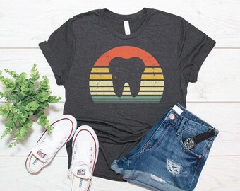 Puede incluir: Una camiseta de manga corta en color gris carbón oscuro con un gráfico central de la silueta de un diente sobre un diseño de atardecer semicircular, con rayas horizontales en tonos naranja, amarillo y verde azulado con textura desgastada.