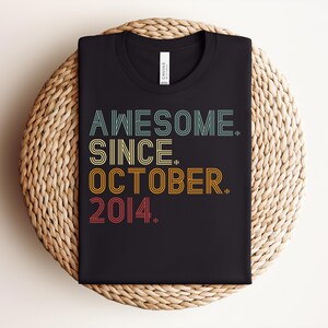 Puede incluir: Una camiseta negra de manga corta con el texto AWESOME. SINCE. OCTOBER. 2014. impreso en una fuente de estilo retro de varias l&iacute;neas con detalles de l&iacute;neas horizontales. Los colores del texto son azul claro, amarillo, naranja y rojo.