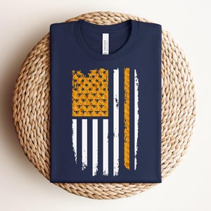 May include: A navy blue t-shirt featuring a graphic of the American flag stylized with a honeycomb pattern. The canton area displays a yellow honeycomb texture with small black bees, while the stripes are rendered in white and yellow honeycomb.