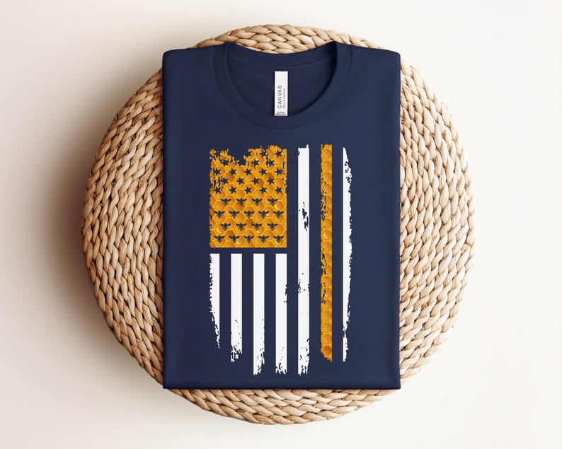 May include: A navy blue t-shirt featuring a graphic of the American flag stylized with a honeycomb pattern. The canton area displays a yellow honeycomb texture with small black bees, while the stripes are rendered in white and yellow honeycomb.