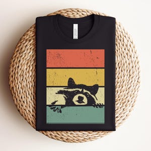 Puede incluir: Camiseta negra con un dise&ntilde;o retro de un mapache asom&aacute;ndose por encima de un fondo a rayas de colores. Las rayas son de color naranja, amarillo y turquesa. La ilustraci&oacute;n del mapache tiene un aspecto vintage.