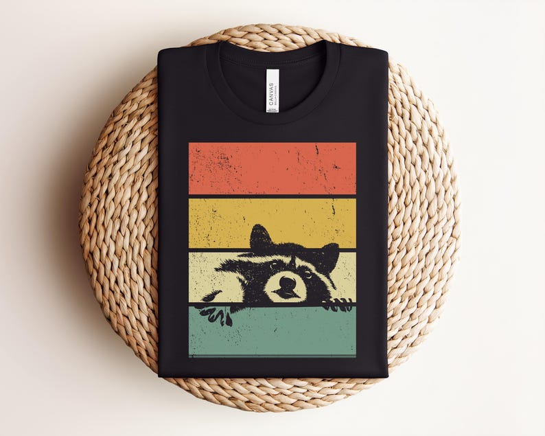 Puede incluir: Camiseta negra con un dise&ntilde;o retro de un mapache asom&aacute;ndose por encima de un fondo a rayas de colores. Las rayas son de color naranja, amarillo y turquesa. La ilustraci&oacute;n del mapache tiene un aspecto vintage.