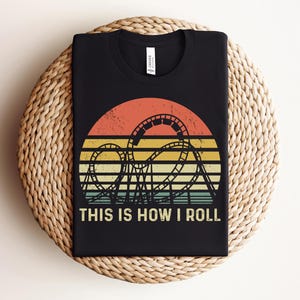 Może przedstawiać: Czarny t-shirt z retro wzorem kolejki g&oacute;rskiej. Grafika przedstawia sylwetkę kolejki g&oacute;rskiej na tle zachodu słońca w kolorach pomarańczowym, ż&oacute;łtym i zielonym. Tekst "THIS IS HOW I ROLL" jest wydrukowany na biało. T-shirt jest prezentowany na tkanej macie.
