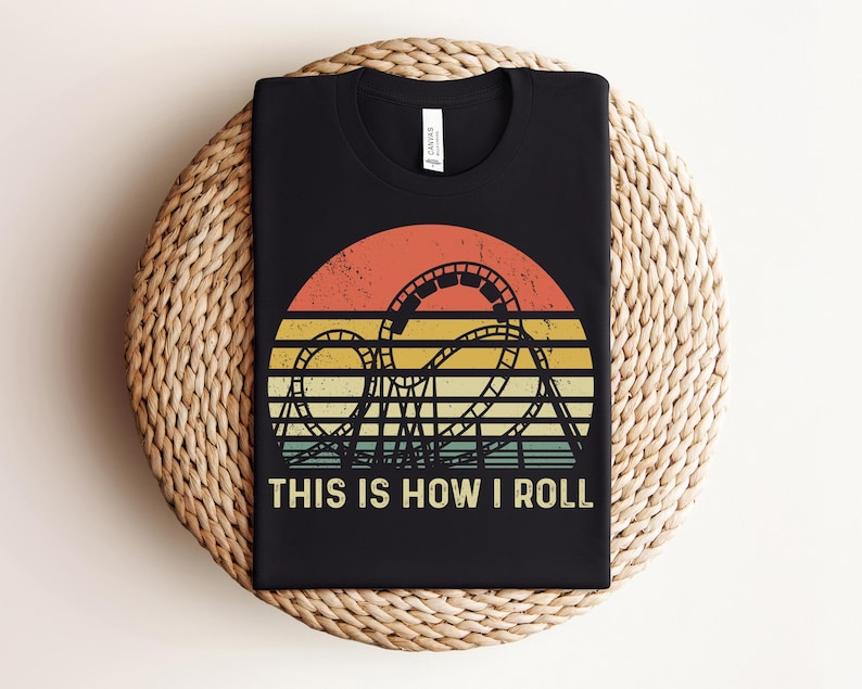 Może przedstawiać: Czarny t-shirt z retro wzorem kolejki g&oacute;rskiej. Grafika przedstawia sylwetkę kolejki g&oacute;rskiej na tle zachodu słońca w kolorach pomarańczowym, ż&oacute;łtym i zielonym. Tekst "THIS IS HOW I ROLL" jest wydrukowany na biało. T-shirt jest prezentowany na tkanej macie.