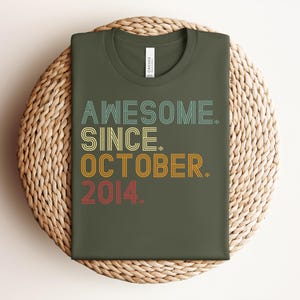 Puede incluir: Una camiseta de cuello redondo color verde oliva con el texto "AWESOME. SINCE. OCTOBER. 2014." impreso en un estilo de fuente retro de varias l&iacute;neas. La palabra "AWESOME." es azul claro, "SINCE." es amarilla, "OCTOBER." es naranja y "2014." es roja.