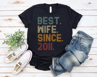 Puede incluir: Una camiseta negra de manga corta con el texto "BEST. WIFE. SINCE. 2011." en una fuente de líneas superpuestas multicolor. La camiseta se muestra junto a unos vaqueros azules y zapatillas de lona blancas, con una pequeña planta verde.