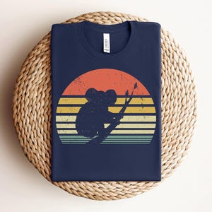 May include: A navy blue t-shirt with a vintage-style sunset graphic featuring a koala bear sitting on a branch. The graphic has a faded, retro look with orange, yellow, and red colors.