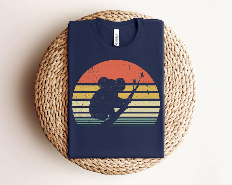 May include: A navy blue t-shirt with a vintage-style sunset graphic featuring a koala bear sitting on a branch. The graphic has a faded, retro look with orange, yellow, and red colors.