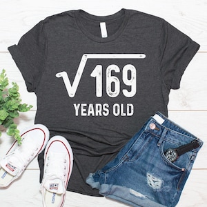 May include: A dark charcoal gray short-sleeve t-shirt featuring a white graphic of a square root symbol over the number 169, followed by the text "YEARS OLD" in a bold, distressed white font centered on the chest of the garment.