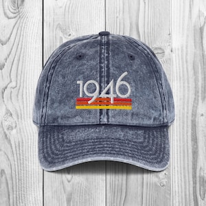 May include: A denim blue baseball cap with the year "1946" embroidered in white. Below the numbers are three horizontal stripes in red, yellow, and orange. The cap has a curved brim and a distressed look.