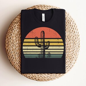 May include: Black t-shirt with a retro sunset graphic featuring a tall cactus in the center. The sunset is in shades of orange, yellow, and blue.