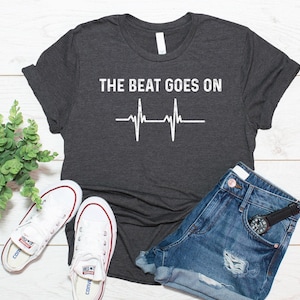 May include: A dark charcoal grey short-sleeve t-shirt featuring the white text THE BEAT GOES ON above a white EKG heart rhythm line graphic. The shirt is displayed alongside a pair of white canvas trainers and blue denim shorts.