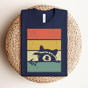 Puede incluir: Camiseta azul marino con un dise&ntilde;o retro. Presenta un mapache asom&aacute;ndose por detr&aacute;s de rayas horizontales naranjas, amarillas y verdes. La camiseta est&aacute; doblada sobre una superficie tejida.