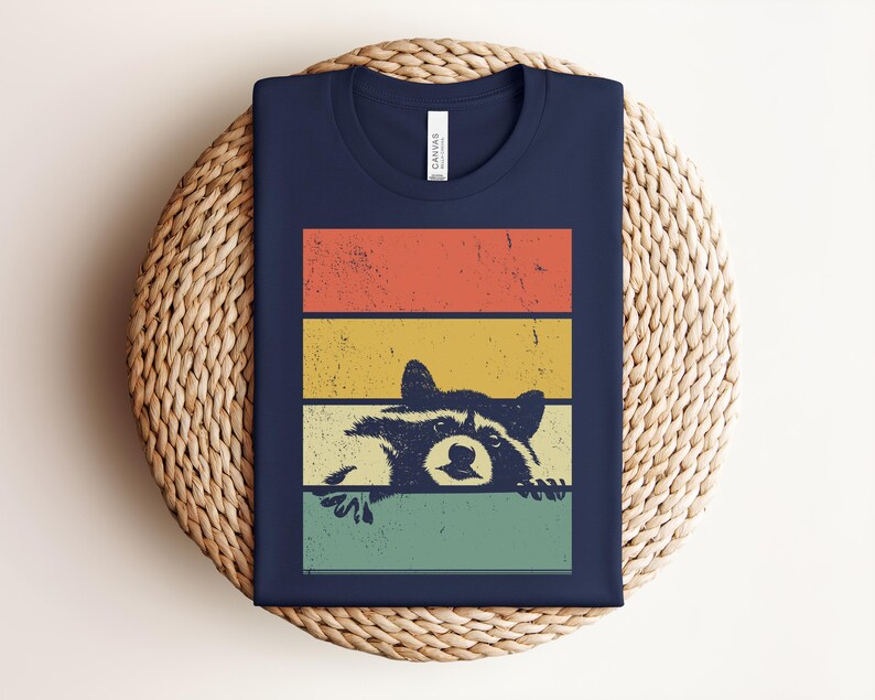 Puede incluir: Camiseta azul marino con un dise&ntilde;o retro. Presenta un mapache asom&aacute;ndose por detr&aacute;s de rayas horizontales naranjas, amarillas y verdes. La camiseta est&aacute; doblada sobre una superficie tejida.