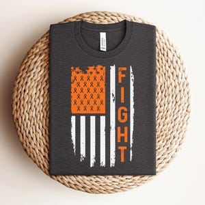 Könnte beinhalten: Ein dunkelgraues T-Shirt mit einem Flaggendesign aus orangefarbenen und weißen Streifen. Der orangefarbene Bereich zeigt ein Muster aus kleinen Awareness-Schleifen. Das Wort FIGHT ist vertikal in orangefarbenen Buchstaben rechts aufgedruckt.