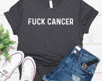 Puede incluir: Una camiseta de manga corta en color gris carbón oscuro con el texto blanco desgastado FUCK CANCER impreso en el pecho. La camiseta se muestra junto a unos pantalones cortos de mezclilla azul, un reloj de pulsera negro y zapatillas blancas.