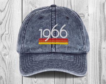 May include: A dark blue acid-wash baseball cap featuring the embroidered white text 1966 above three horizontal stripes in red, orange, and yellow. The cap has a curved brim and a six-panel construction with a button at the top centre.
