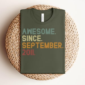 May include: An olive green t-shirt featuring the text "AWESOME. SINCE. SEPTEMBER. 2011." in a retro, multi-colored, layered font style. The shirt has a crew neckline and a white brand label visible at the inner collar area.