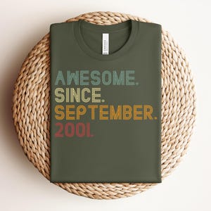 May include: An olive green t-shirt featuring the text "AWESOME. SINCE. SEPTEMBER. 2001." in a multi-colored, retro-style layered font. The word "AWESOME." is in light blue, "SINCE." is in yellow, "SEPTEMBER." is in orange, and "2001." is in red.