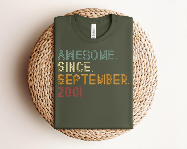 May include: An olive green t-shirt featuring the text "AWESOME. SINCE. SEPTEMBER. 2001." in a multi-colored, retro-style layered font. The word "AWESOME." is in light blue, "SINCE." is in yellow, "SEPTEMBER." is in orange, and "2001." is in red.