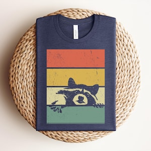 Puede incluir: Camiseta azul marino con un dise&ntilde;o retro de mapache, asom&aacute;ndose por encima de un bloque de color naranja, amarillo y verde. La camiseta est&aacute; doblada sobre una superficie tejida.