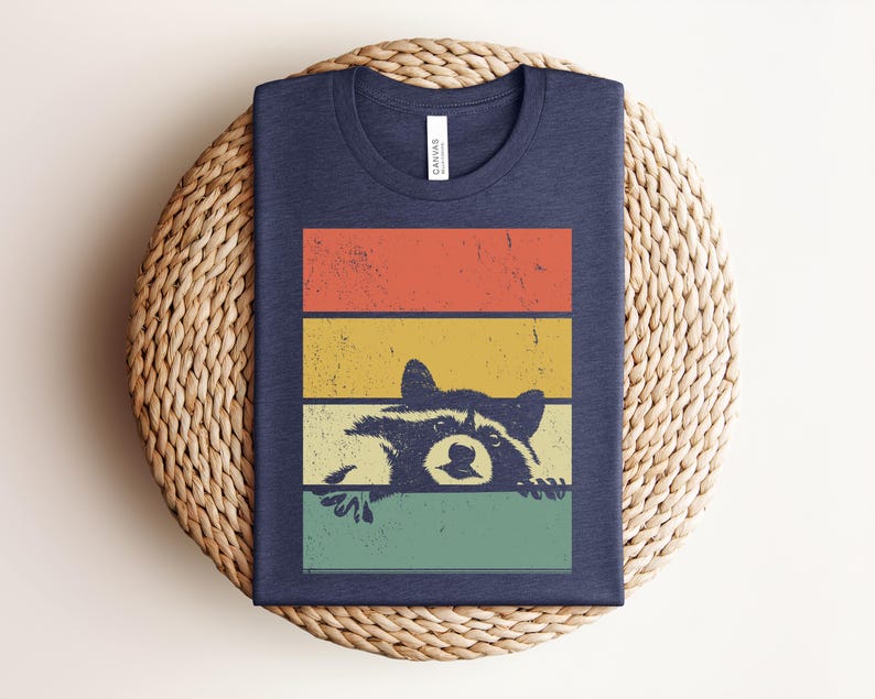 Puede incluir: Camiseta azul marino con un dise&ntilde;o retro de mapache, asom&aacute;ndose por encima de un bloque de color naranja, amarillo y verde. La camiseta est&aacute; doblada sobre una superficie tejida.