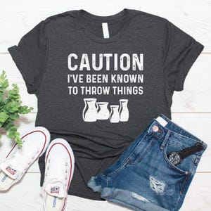 May include: A dark heather gray short-sleeve t-shirt featuring the white distressed text CAUTION I'VE BEEN KNOWN TO THROW THINGS above a graphic of four pottery vases. The shirt is paired with blue denim shorts and white canvas sneakers.