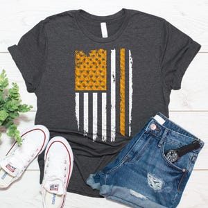 May include: A dark charcoal gray short-sleeve t-shirt featuring a graphic of the American flag. The flag design incorporates a yellow honeycomb pattern with small black bees in the canton and a vertical yellow honeycomb stripe.