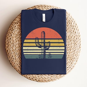 May include: A navy blue t-shirt with a retro sunset graphic featuring a silhouette of a cactus. The sunset is in shades of orange, yellow, and blue.
