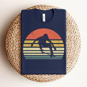 May include: A navy blue t-shirt with a vintage-style graphic of a person skateboarding in front of a sunset. The graphic has a faded, retro look with orange, yellow, and red colors.