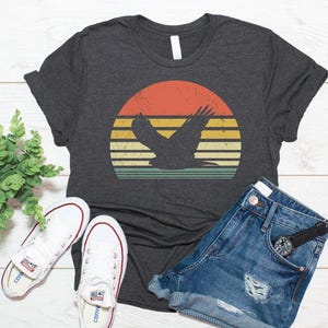 May include: A dark charcoal grey short-sleeve t-shirt featuring a graphic of a bird in flight silhouetted against a circular sunset design with horizontal stripes in shades of orange, yellow, and teal, paired with white canvas trainers.