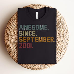 May include: A black t-shirt featuring the text "AWESOME. SINCE. SEPTEMBER. 2001." in a multi-colored, retro-inspired font. The words are stacked vertically, with each line in a different color including teal, yellow, orange, and red.