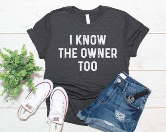 Może przedstawiać: Ciemnoszary melanżowy t-shirt z krótkim rękawem z białym, postarzanym napisem "I KNOW THE OWNER TOO" nadrukowanym z przodu. Koszulka jest zestawiona z niebieskimi szortami dżinsowymi, czarnym zegarkiem na rękę i białymi trampkami.