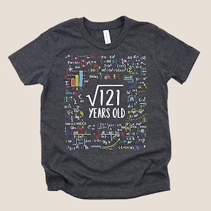 May include: A dark heather gray short-sleeve t-shirt featuring a white graphic in the center that reads square root of 121 YEARS OLD. The design is surrounded by various colorful mathematical formulas, symbols, and equations in white, red, blue, and yellow.