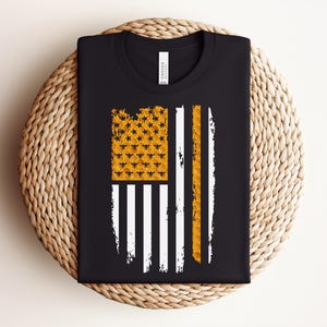 May include: A black crew neck t-shirt featuring a graphic of a stylised American flag. The flag's canton is filled with a yellow honeycomb pattern containing small black bee silhouettes, while the stripes are a mix of white and yellow honeycomb.