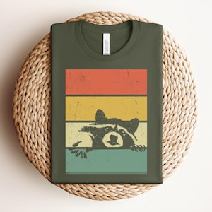 Puede incluir: Camiseta verde oliva con un dise&ntilde;o retro. Presenta una ilustraci&oacute;n de un mapache asom&aacute;ndose por encima de un fondo a rayas rojas, amarillas y turquesas. El dise&ntilde;o tiene una est&eacute;tica vintage y desgastada.