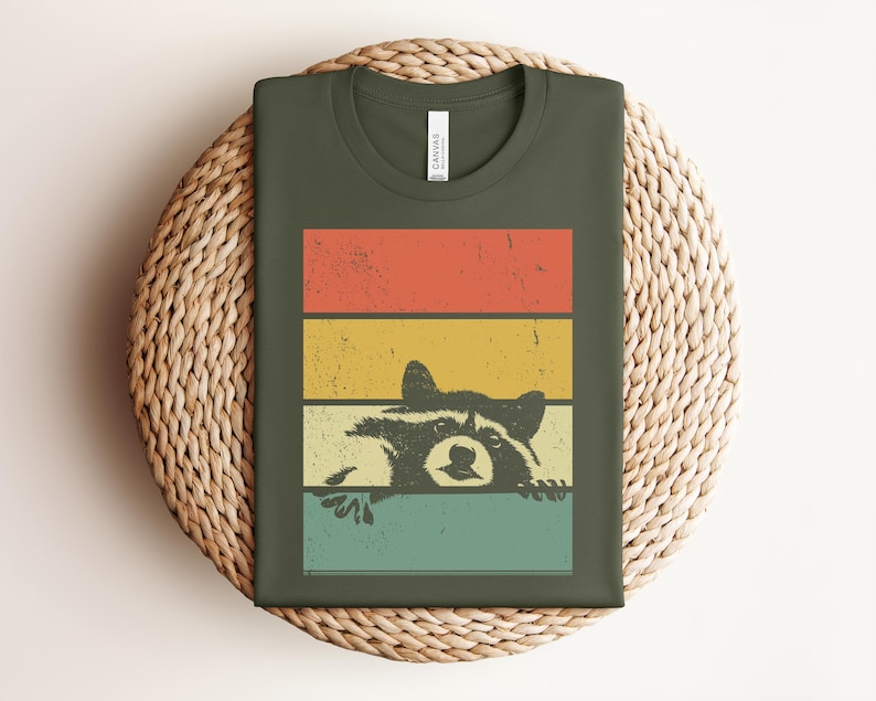 Puede incluir: Camiseta verde oliva con un dise&ntilde;o retro. Presenta una ilustraci&oacute;n de un mapache asom&aacute;ndose por encima de un fondo a rayas rojas, amarillas y turquesas. El dise&ntilde;o tiene una est&eacute;tica vintage y desgastada.