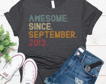 May include: A dark charcoal gray short-sleeve t-shirt featuring the text AWESOME. SINCE. SEPTEMBER. 2013. in a retro-inspired, multi-colored layered font with teal, yellow, orange, and red stripes against the dark fabric background.
