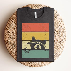 Puede incluir: Camiseta gris oscuro con un dise&ntilde;o retro que presenta un mapache asom&aacute;ndose sobre un fondo de rayas de colores. Las rayas son rojas, amarillas y turquesas. La camiseta est&aacute; doblada y tiene una etiqueta en el cuello.