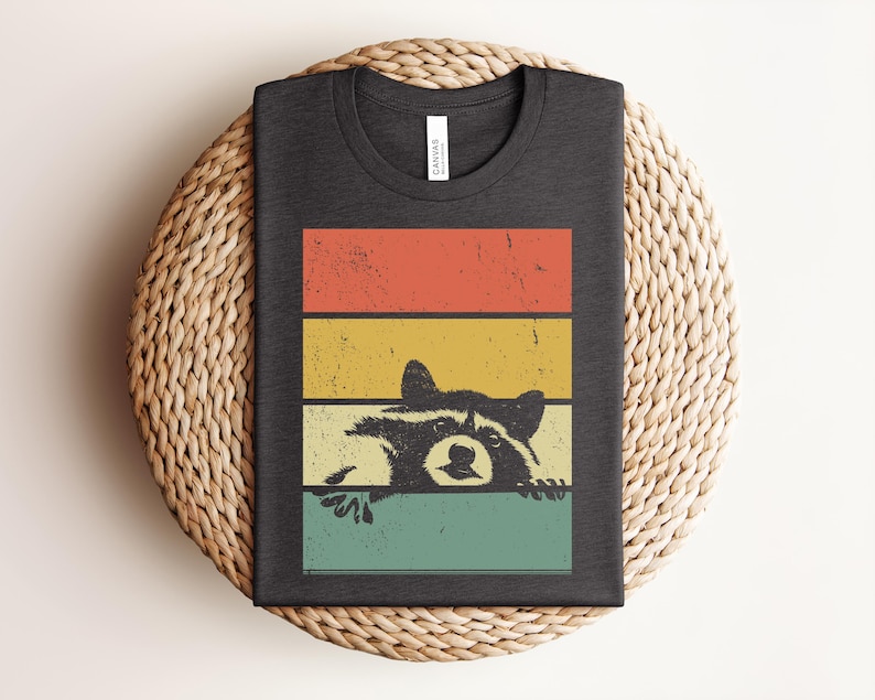 Puede incluir: Camiseta gris oscuro con un dise&ntilde;o retro que presenta un mapache asom&aacute;ndose sobre un fondo de rayas de colores. Las rayas son rojas, amarillas y turquesas. La camiseta est&aacute; doblada y tiene una etiqueta en el cuello.