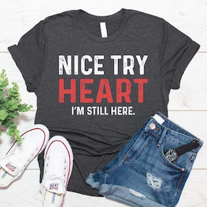 Pode incluir: Uma t-shirt de manga curta em cinzento-carvão escuro com o texto branco e vermelho "NICE TRY HEART I'M STILL HERE." A t-shirt é apresentada com calções de ganga azuis, ténis de lona brancos e um relógio de pulso preto com mostrador prateado.